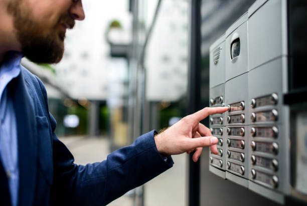 A man in a suit presses a button on an intercom system outside a building.