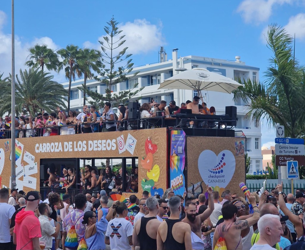 A vibrant scene from the Maspalomas Pride parade, featuring a decorated float with partygoers and spectators celebrating in the street.
