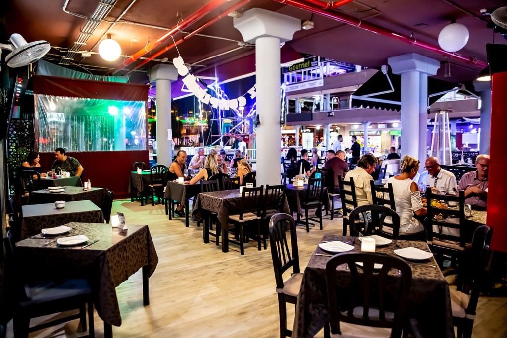 A lively dining area in a bar or restaurant in Maspalomas, Gran Canaria, with dark tables and chairs, people enjoying meals and drinks, and colorful lights in the background.
